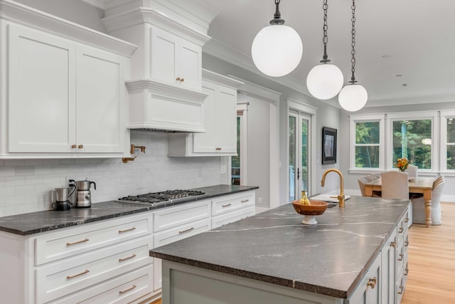 kitchen featuring light wood-type flooring, a center island with sink, stainless steel gas stovetop, backsplash, and pendant lighting