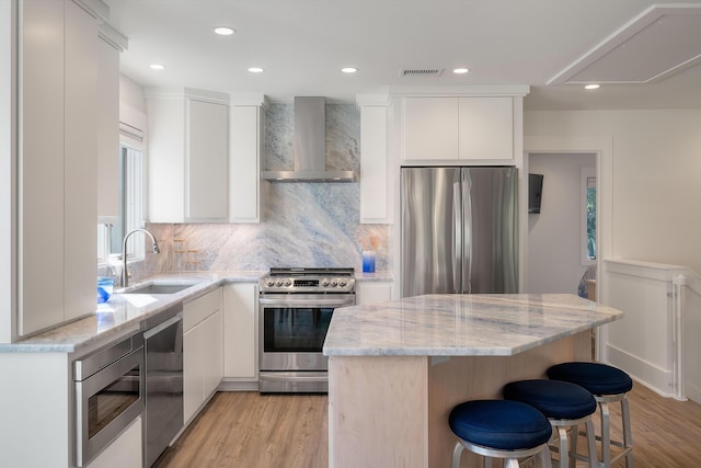 kitchen featuring wall chimney range hood, a sink, a center island, stainless steel appliances, and light wood-type flooring