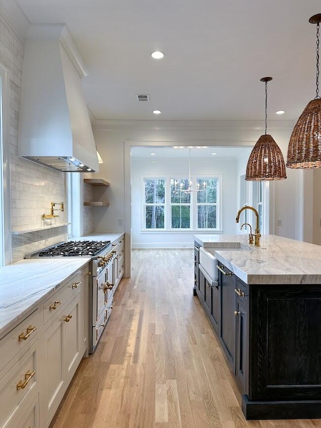 kitchen featuring a kitchen island with sink, wall chimney range hood, hanging light fixtures, sink, and stainless steel range