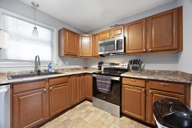 kitchen featuring brown cabinets, a sink, stone finish flooring, appliances with stainless steel finishes, and hanging light fixtures