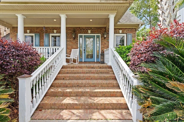 doorway to property with brick siding, a porch, and a shingled roof