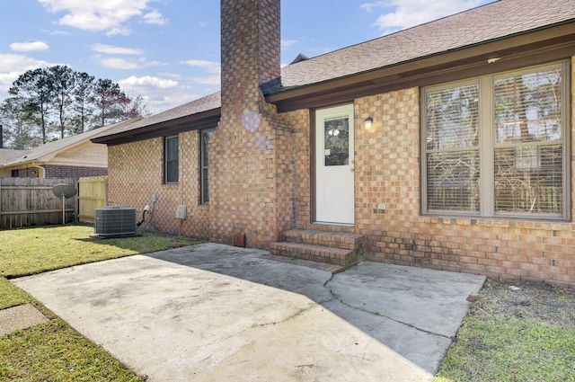 view of exterior entry with brick siding, fence, a lawn, a chimney, and a patio