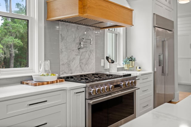 kitchen with premium appliances, white cabinetry, custom range hood, backsplash, and light stone countertops