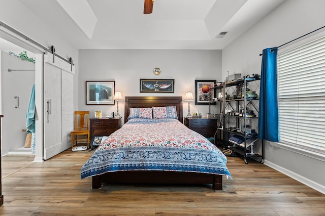 bedroom with ceiling fan, a tray ceiling, a barn door, and light hardwood / wood-style flooring