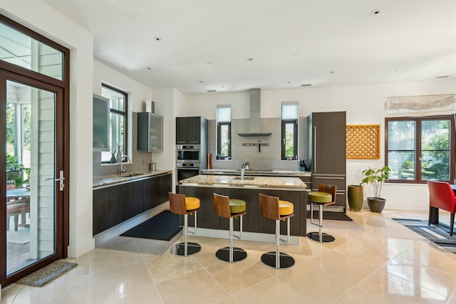 kitchen with wall chimney exhaust hood, a healthy amount of sunlight, a kitchen island with sink, light stone counters, and decorative backsplash