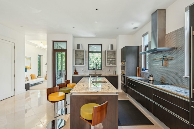 kitchen featuring backsplash, light stone counters, wall chimney range hood, a center island with sink, and a chandelier