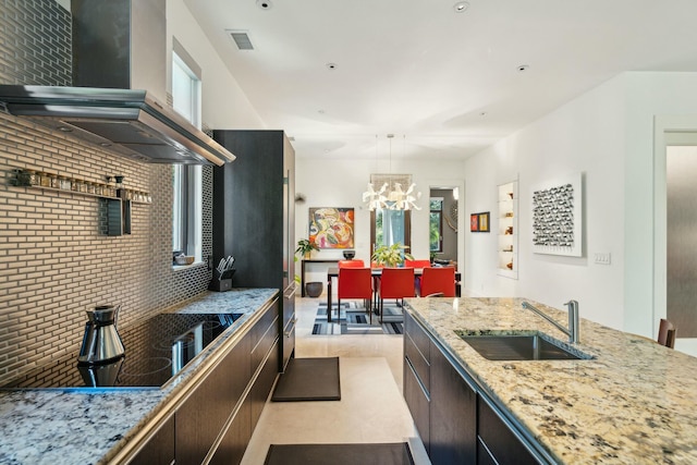 kitchen with light stone countertops, sink, wall chimney range hood, a notable chandelier, and dark brown cabinets