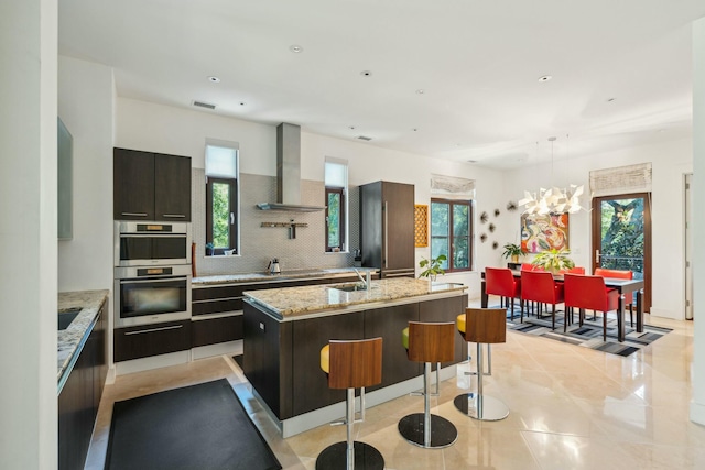 kitchen featuring pendant lighting, an inviting chandelier, a center island with sink, wall chimney exhaust hood, and a wealth of natural light