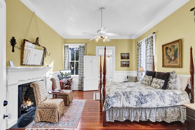 bedroom featuring a wainscoted wall, a decorative wall, ornamental molding, a warm lit fireplace, and hardwood / wood-style flooring