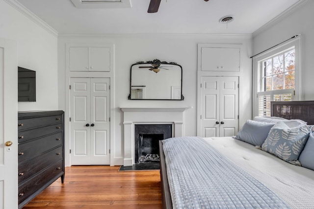 bedroom with crown molding, visible vents, a fireplace with flush hearth, ceiling fan, and wood finished floors