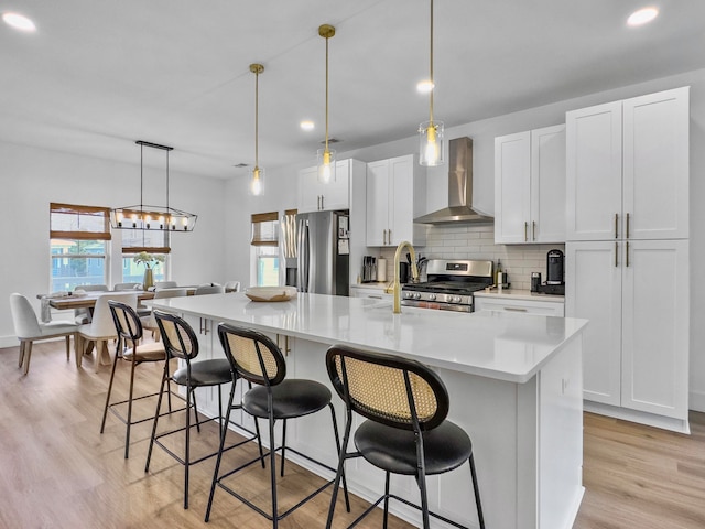 kitchen featuring light wood-type flooring, wall chimney exhaust hood, stainless steel appliances, a kitchen island with sink, and white cabinetry