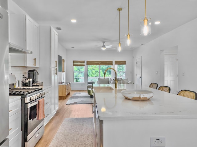 kitchen featuring stainless steel gas range oven, pendant lighting, light hardwood / wood-style flooring, white cabinets, and an island with sink