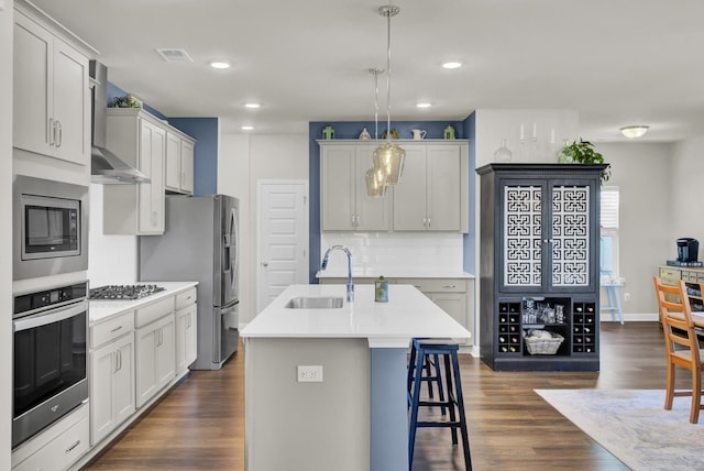 kitchen featuring sink, pendant lighting, wall chimney range hood, stainless steel appliances, and an island with sink