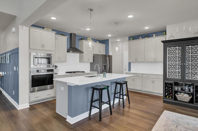 kitchen featuring a center island with sink, hanging light fixtures, white cabinets, appliances with stainless steel finishes, and wall chimney range hood