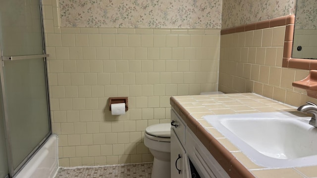bathroom featuring tile walls, vanity, bath / shower combo with glass door, toilet, and wallpapered walls