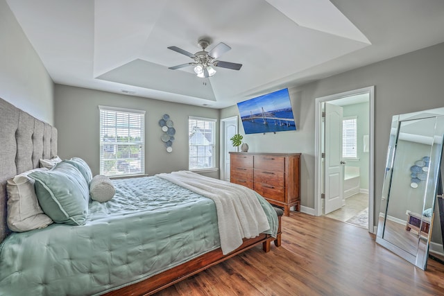 bedroom featuring baseboards, ensuite bath, a tray ceiling, ceiling fan, and wood finished floors