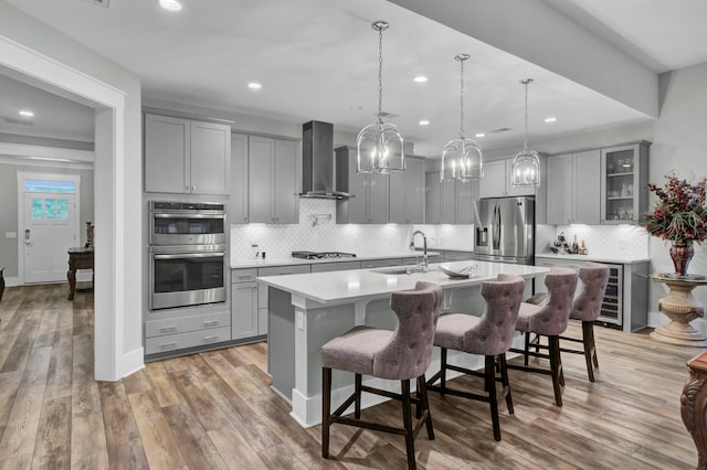 kitchen featuring a center island with sink, wall chimney exhaust hood, hardwood / wood-style flooring, pendant lighting, and appliances with stainless steel finishes