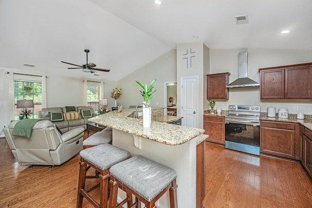 kitchen featuring wall chimney exhaust hood, a breakfast bar, wood-type flooring, a center island with sink, and stainless steel electric range