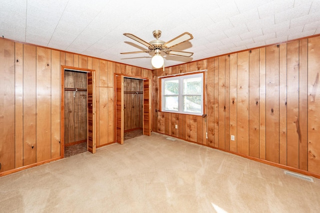 unfurnished bedroom with ceiling fan, wood walls, light carpet, and multiple closets