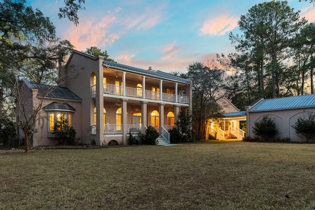 back house at dusk with a balcony, covered porch, and a yard