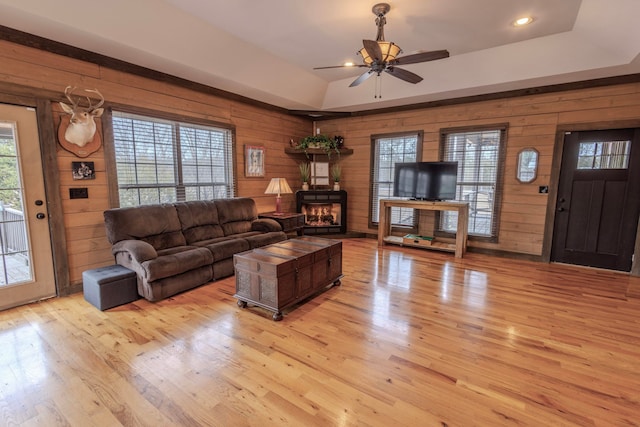 living room with a tray ceiling, wood walls, light hardwood / wood-style floors, and ceiling fan