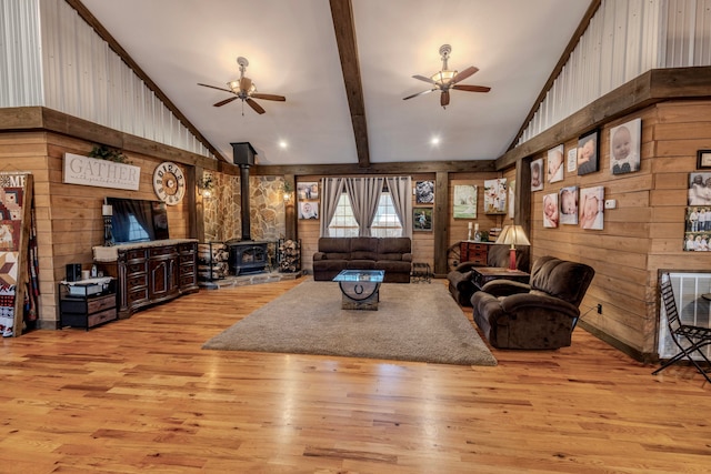 living room with ceiling fan, a wood stove, light wood-type flooring, and vaulted ceiling with beams