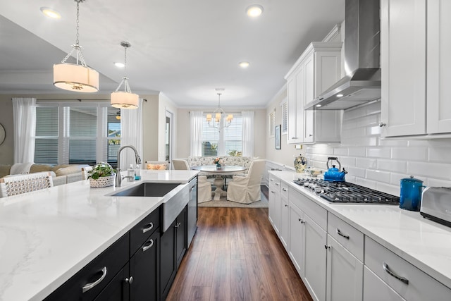 kitchen with wall chimney range hood, sink, hanging light fixtures, light stone countertops, and white cabinets