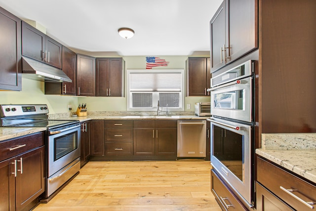 kitchen featuring appliances with stainless steel finishes, a sink, light stone countertops, light wood-type flooring, and under cabinet range hood