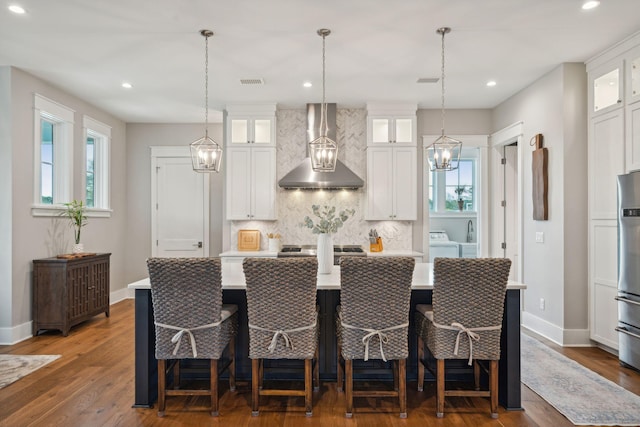 kitchen with light countertops, glass insert cabinets, white cabinets, wall chimney range hood, and an island with sink