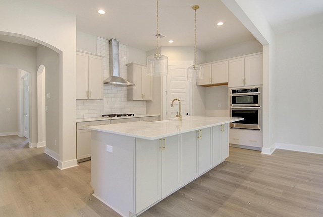 kitchen with arched walkways, double oven, light wood-style floors, wall chimney exhaust hood, and tasteful backsplash