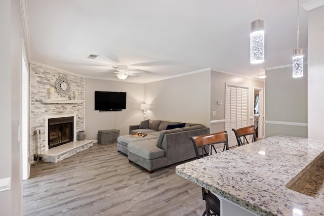 living area featuring a ceiling fan, visible vents, light wood-style flooring, ornamental molding, and a stone fireplace