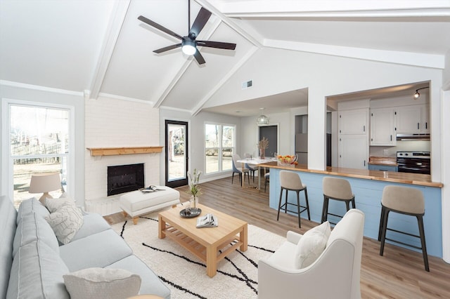 living room with light wood-type flooring, vaulted ceiling with beams, a brick fireplace, and ceiling fan