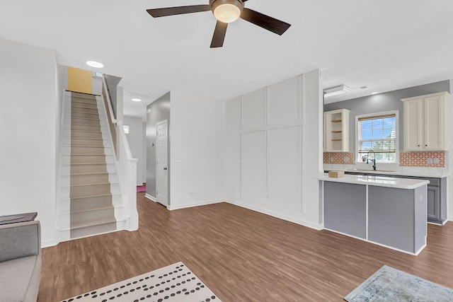 unfurnished living room with a sink, wood finished floors, ceiling fan, stairway, and baseboards