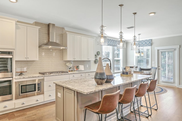 kitchen with wall chimney exhaust hood, stainless steel appliances, pendant lighting, light hardwood / wood-style flooring, and an island with sink