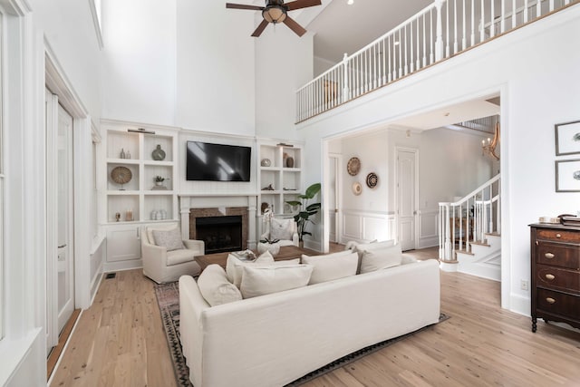 living area featuring stairs, ceiling fan with notable chandelier, a fireplace, a high ceiling, and light wood-style floors