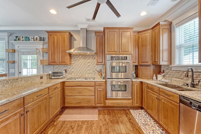 kitchen with appliances with stainless steel finishes, wall chimney exhaust hood, a sink, a ceiling fan, and light wood-style floors
