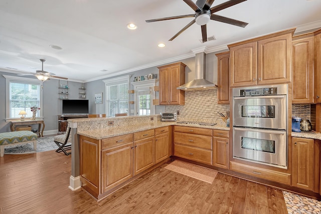 kitchen featuring stainless steel appliances, ceiling fan, wall chimney range hood, a peninsula, and crown molding