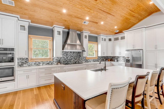 kitchen with sink, wall chimney exhaust hood, a kitchen island with sink, wood ceiling, and appliances with stainless steel finishes