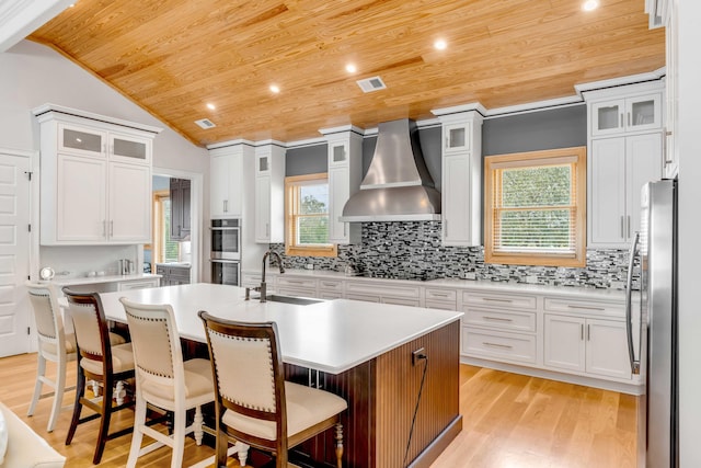 kitchen featuring wall chimney exhaust hood, sink, a center island with sink, wooden ceiling, and white cabinetry