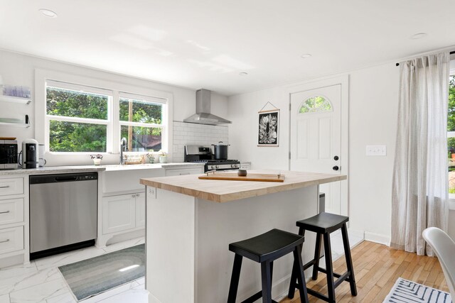 kitchen featuring stainless steel appliances, wall chimney exhaust hood, butcher block countertops, a breakfast bar area, and white cabinetry