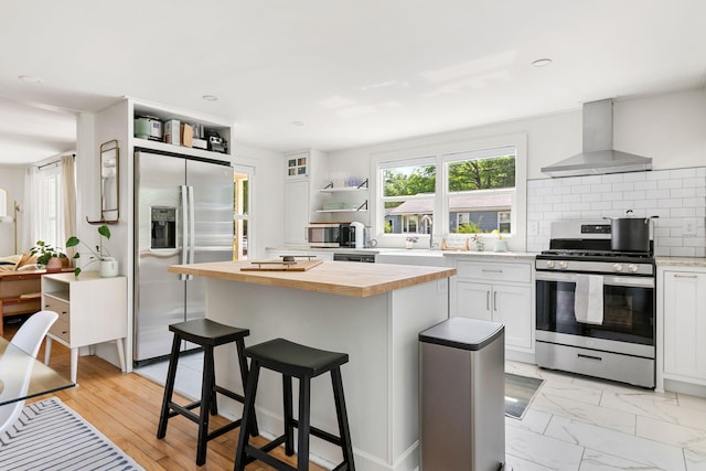 kitchen with appliances with stainless steel finishes, wall chimney range hood, wood counters, open shelves, and white cabinets