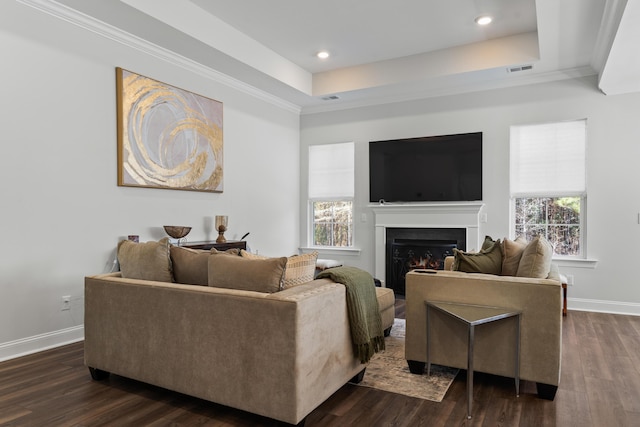 living room featuring baseboards, a tray ceiling, dark wood-style flooring, a warm lit fireplace, and crown molding