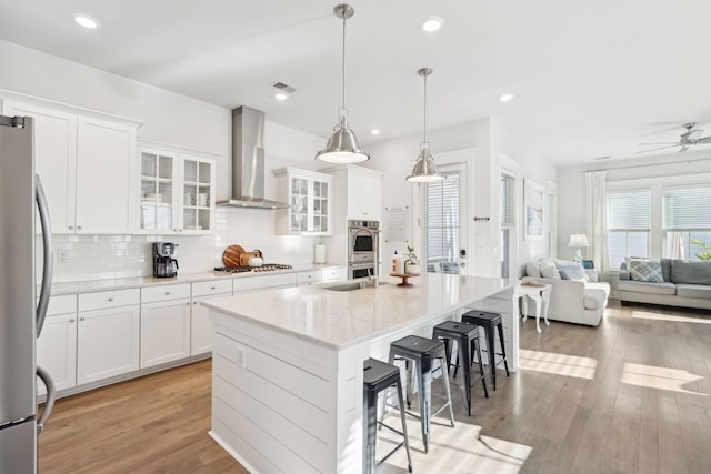 kitchen featuring white cabinetry, wall chimney range hood, an island with sink, and appliances with stainless steel finishes