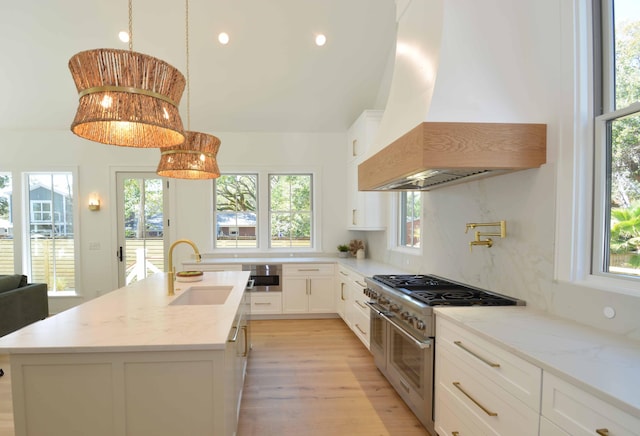 kitchen with a center island with sink, a sink, white cabinetry, range with two ovens, and custom exhaust hood