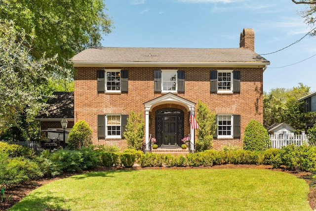 colonial home featuring fence, a shingled roof, a front lawn, brick siding, and a chimney