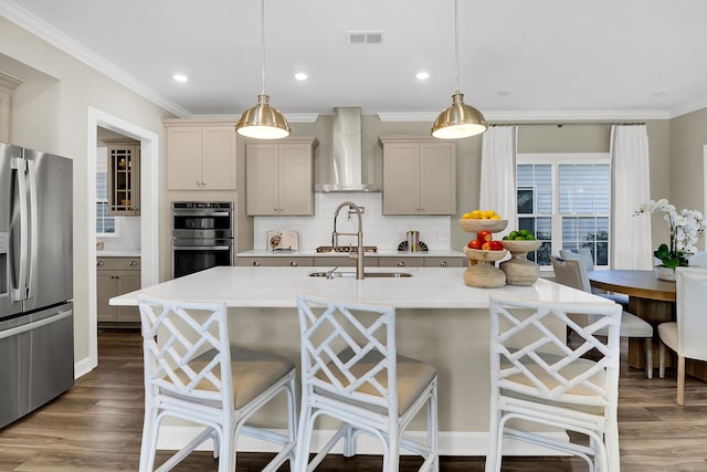 kitchen featuring pendant lighting, wall chimney range hood, appliances with stainless steel finishes, a kitchen island with sink, and a kitchen bar