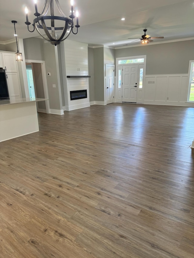 unfurnished living room featuring ceiling fan with notable chandelier, a large fireplace, crown molding, and dark wood-type flooring