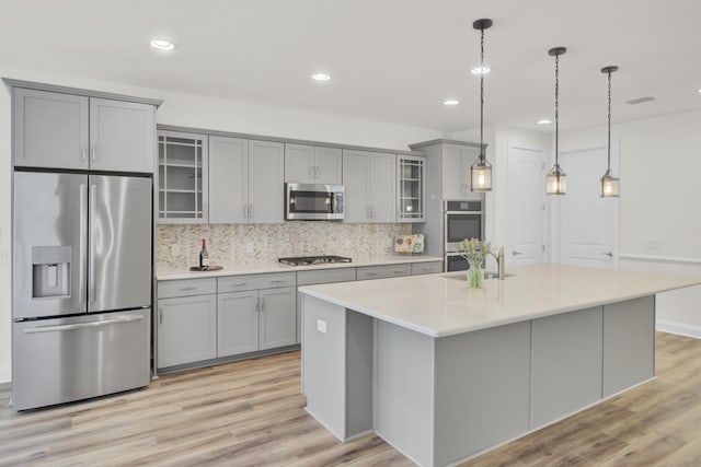 kitchen with stainless steel appliances, a center island with sink, light wood-type flooring, backsplash, and gray cabinets
