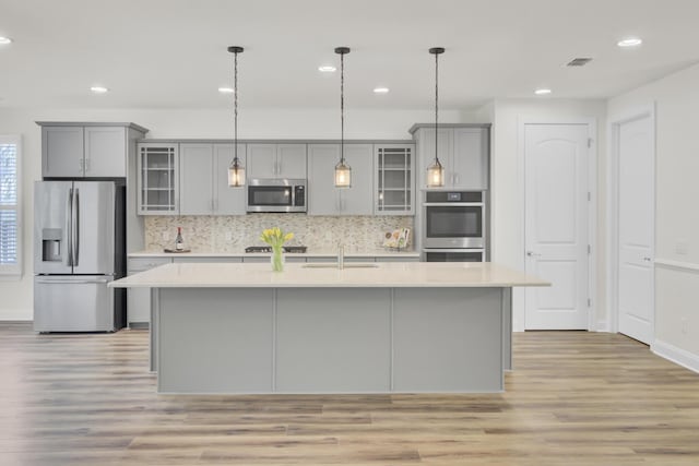 kitchen with a sink, an island with sink, gray cabinets, light countertops, and appliances with stainless steel finishes