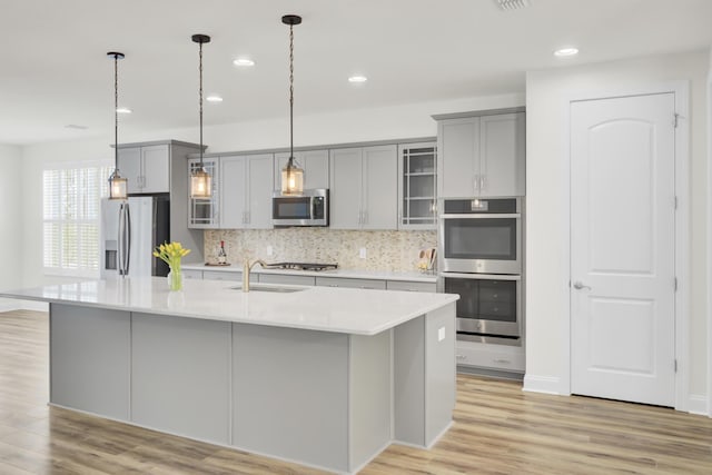 kitchen with gray cabinets, stainless steel appliances, a kitchen island with sink, and light wood-style flooring
